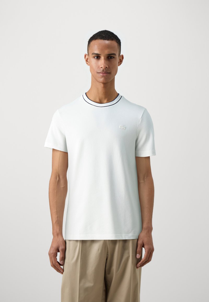 T-shirt bianco a maniche corte con colletto a coste, caratterizzato da una striscia blu navy a contrasto e un logo ricamato sul lato sinistro del petto.