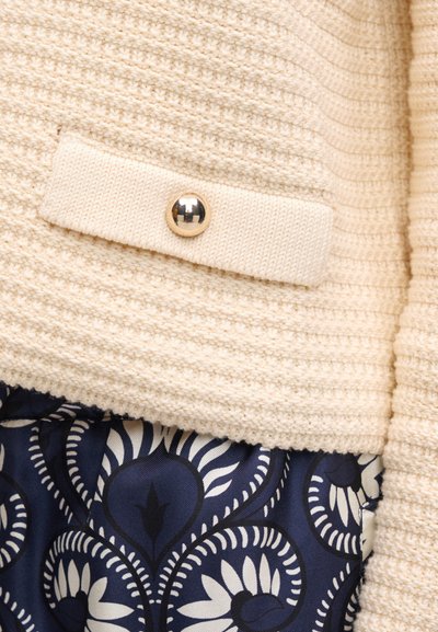 Πλεκτό ύφασμα εκρού με ένα μικρό καπάκι που διαθέτει στρογγυλό μεταλλικό κουμπί πάνω από ύφασμα με μοτίβο σκοτεινού μπλε και λευκά λουλούδια.
