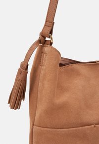 Borsa in suede marrone con una consistenza morbida, dotata di una tracolla intrecciata e di un pon-pon decorativo. Fodera interna liscia, design minimalista.