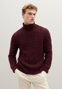 Weinroter Zopfmusterpullover mit hohem Kragen und gerippten Bündchen, getragen über cremefarbenen Hosen, mit strukturierten, dicken Garnmustern.