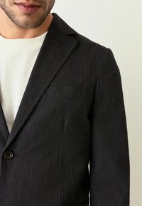 Zbliżenie na mężczyznę w ciemnej marynarce w prążki, noszącym jasny sweter, z wyróżnieniem detali guzików i kieszeni marynarki.
