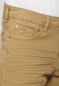 Pantalons khaki présentés en gros plan. Fabriqués en tissu denim, avec une coupe slim, des coutures visibles, une poche avant et une étiquette en cuir avec une marque.