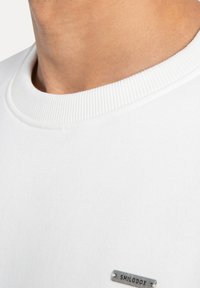 Sudadera blanca de cuello redondo hecha de algodón suave. Presenta un cuello acanalado y una pequeña etiqueta de logotipo metálico en la parte inferior derecha. Textura suave.