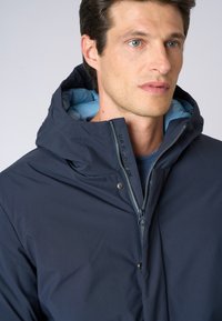 Giacca a vento blu scuro con chiusura a zip e fodera contrastante blu chiaro. Presenta uno snap anteriore, una texture liscia e un design minimalista.