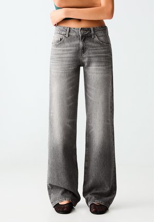 Personne portant un jean gris délavé taille haute à jambes larges, associé à des chaussures noires en mesh, debout avec les bras croisés sur le ventre découvert.
