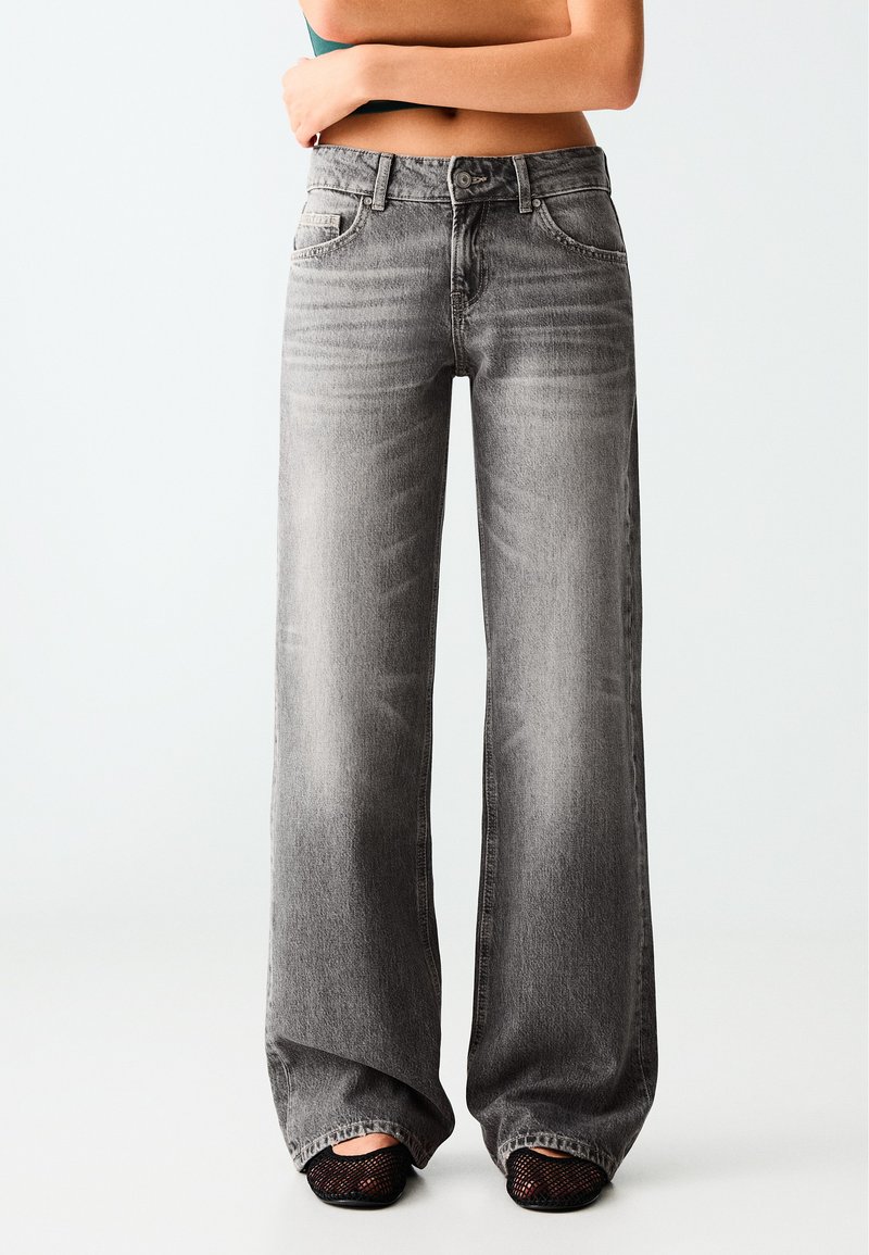 Personne portant un jean gris délavé taille haute à jambes larges, associé à des chaussures noires en mesh, debout avec les bras croisés sur le ventre découvert.