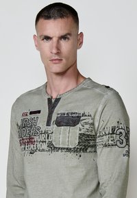 Hosszú ujjú olíva zöld ing kontrasztos varrással, mellzsebbel, grafikus mintákkal és gombos díszítéssel a nyakvonalon.