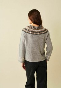 Pitkät ruskeat hiukset omaava nainen, joka pukeutuu harmaaseen, kuvioidulla yläosalla varustettuun villapaitaan ja mustiin housuihin, seisoo yksivärisen beigen taustan edessä.