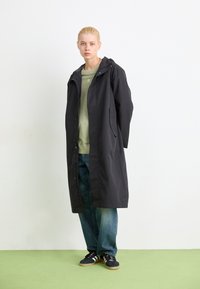 Svart lang regnfrakk med hette, med frontknapplukking og to side lommer, brukt over en grønn skjorte og blå jeans.