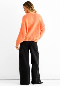 Coral knitted sweater with oversized sleeves and ribbed hem, paired with high-waisted black wide-leg jeans. Smooth textured fabric.