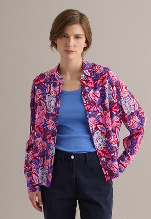 Mujer que lleva una camiseta sin mangas azul bajo una camisa abotonada de manga larga con patrones florales en rojo, rosa y azul, combinada con pantalones oscuros.
