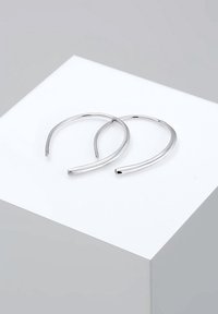 Deux boucles d'oreilles argentées élégantes et ouvertes reposant sur un piédestal géométrique blanc, avec un fond gris.