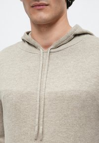 Homme portant un sweat à capuche en maille beige clair avec cordons, visible du cou au milieu du torse sur un fond blanc uni.