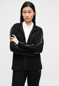 Zwarte jas met ritssluiting en hoge kraag, met reflecterende accenten langs de mouwen. Draagt over een wit poloshirt, gladde textuur.