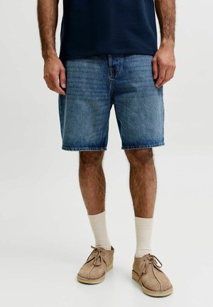 Man met blauwe denimshorts, beige suède veterschoenen, witte sokken en een marineblauw overhemd, staand tegen een effen achtergrond.