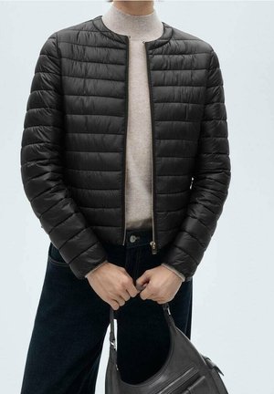 Person trägt eine schwarze gesteppte Jacke über einem beigen Rollkragenpullover, dunkle Hose, hält eine schwarze Lederhandtasche mit beiden Händen vor sich.