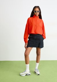 Lys oransje vindjakke, svart snorliv skjørt, hvite sokker og grå joggesko. Modellen har på seg stilige solbriller; glatte teksturer og moderne design.