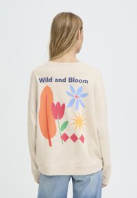 Sweat-shirt beige avec un motif graphique coloré au dos, comprenant des fleurs, des feuilles et des formes. Coupe ample avec poignets côtelés.