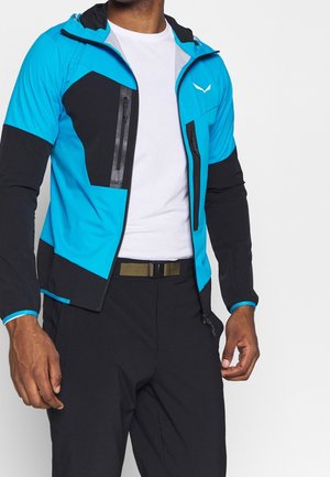 Mann trägt eine blau-schwarze Reißverschlussjacke über einem weißen Hemd und schwarzen Hosen mit einem grünen Gürtel, steht vor einem schlichten Hintergrund.