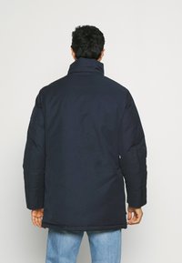 Marinblå puffjacka med hög krage, långa ärmar och en strömlinjeformad silhuett. Tillverkad av hållbart, slätt material med minimal synlig sömnad.