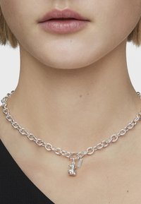 Srebrny łańcuszek z dużymi ogniwami, z małym charms z nied�źwiedziem oraz dodatkowym zawieszką w formie tagu, o gładkiej metalicznej teksturze.