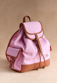Sac à dos en toile rose avec garniture en cuir marron, fermetures à boucle, rabat avant, poche latérale et poignée supérieure sur fond beige.