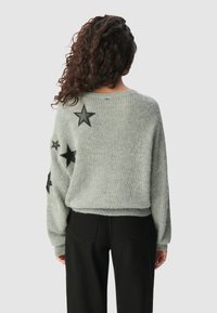 Kobieta z kręconymi włosami, ubrana w szary sweter z dzianiny ozdobiony czarnymi łatkami w kształcie gwiazd i czarne spodnie, zwrócona tyłem na jednolitym tle.