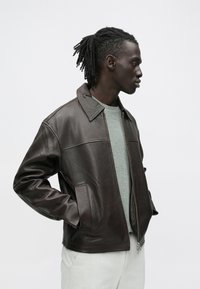 Vīrietis ar dredeļiem, valkājot tumši brūnu ādas jaku pār gaiši pelēku džemperi un gaišām biksēm, stāv profilā ar rokām kabatās.