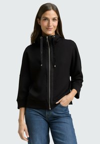 Zwarte hoodie met ritssluiting, hoge kraag, trekkoorddetails en manchetten; gecombineerd met blauwe spijkerbroek en een gladde stoftextuur.