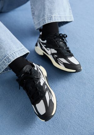 Personne portant des baskets Nike noires et blanches avec des chaussettes noires et un jean bleu clair debout sur un tapis bleu.