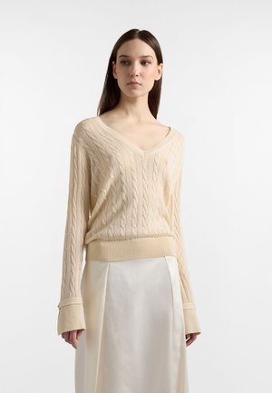 Kvinde iført en cremefarvet kabelstrikket V-hals sweater med lange ærmer og et cremefarvet satinskjørt, stående foran en ensfarvet hvid baggrund.