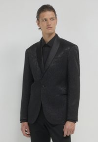 Giacca smoking nera a motivo con rever in raso, chiusura a un bottone e due tasche laterali, indossata sopra una camicia nera. Texture liscia.