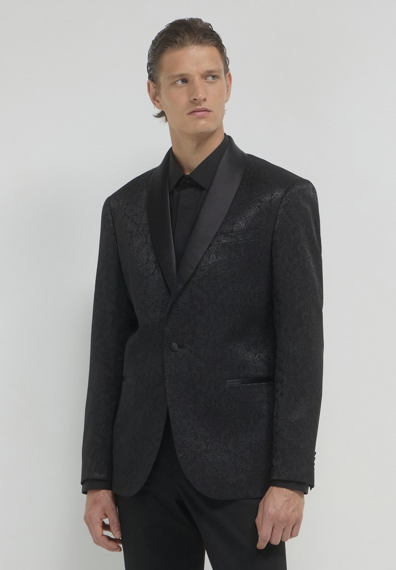 Giacca smoking nera a motivo con rever in raso, chiusura a un bottone e due tasche laterali, indossata sopra una camicia nera. Texture liscia.