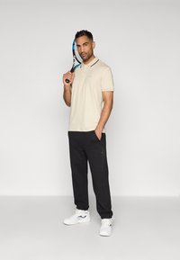 Béžové polo tričko s černým límečkem, doplněné černými tepláky. Bílé tenisky. Osoba drží tenisovou raketu a má neutrální postoj.