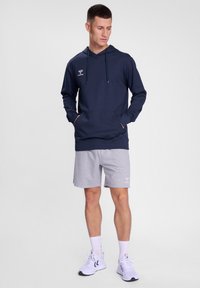 Felpa blu navy con tasca frontale e cappuccio con cordino, abbinata a pantaloni corti grigi. Logo bianco sul lato sinistro del petto e sui pantaloni corti. Abbigliamento sportivo casual.