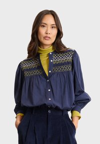 Blusa blu navy con intricati ricami verdi e bianchi sullo scollo, caratterizzata da una chiusura a bottoni e maniche arricciate, abbinata a un dolcevita senape.