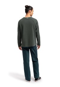 Camicia verde a maniche lunghe con scollatura rotonda e etichetta sul retro, abbinata a pantaloni blu e sneakers nere. Tessuto morbido, vestibilità casual.