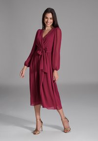 Bordeauxfarbiges Wickelkleid mit langen Ärmeln, tailliertem Bund und fließendem Rock. Glattes, leichtes Material mit dezentem Glanz. Tragen mit goldenen Absatzsandalen.