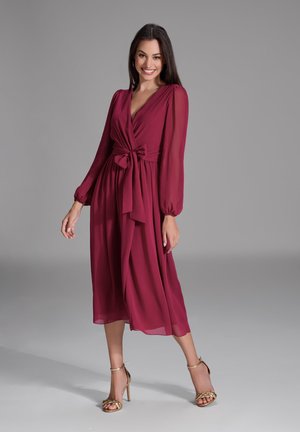 Bordeauxfarbiges Wickelkleid mit langen Ärmeln, tailliertem Bund und fließendem Rock. Glattes, leichtes Material mit dezentem Glanz. Tragen mit goldenen Absatzsandalen.