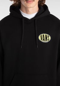 Felpa nera con cappuccio realizzata in tessuto morbido, dotata di tasca frontale. Presenta un logo ovale giallo con scritto "VANS". Include cappuccio regolabile con lacci.
