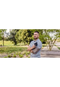 Man in a light gray polo shirt, showcasing a tribal tattoo on his arm, holding a black wallet, with greenery in the background.