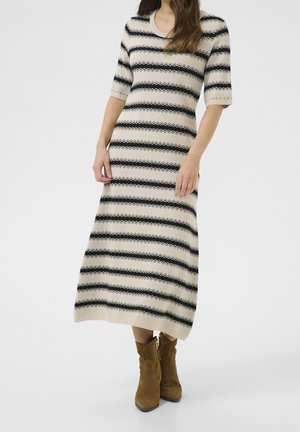 Femme portant une robe midi beige à rayures zigzag noires et manches aux coudes, associée à des bottines marron en daim.