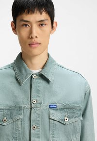 Light blue denim jacket with a collared neck, front button closure, and two chest pockets. Features a branded blue tag on the left side.