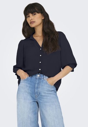 Vrouw met lang donker haar, gekleed in een marineblauw overhemd met knopen en lichtblauwe high-waisted spijkerbroek, met handen in de zakken.