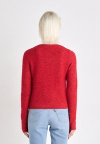 Suéter vermelho justo feito de lã texturizada com mangas longas. Usado com jeans azul claro com bolsos traseiros e costuras subtis.