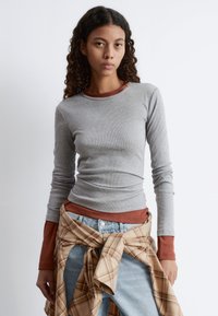 Langärmliges graues geripptes Shirt, das über einem rötlichen Oberteil getragen wird; kombiniert mit hellblauen Jeans und einem beige karierten Hemd, das um die Taille gebunden ist.