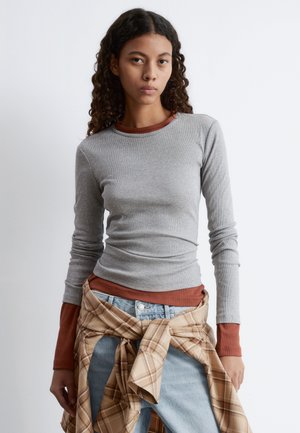 Langärmliges graues geripptes Shirt, das über einem rötlichen Oberteil getragen wird; kombiniert mit hellblauen Jeans und einem beige karierten Hemd, das um die Taille gebunden ist.