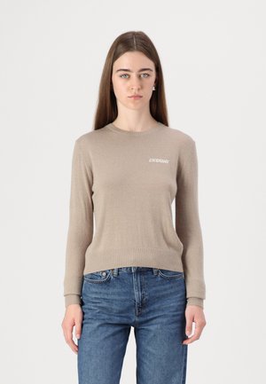 Lys beige strikket sweater med rund hals og lange ærmer, med ribbede manchetter og et lille hvidt logo på brystet. Båret med blå jeans.