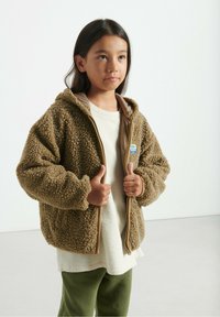 Bambino con capelli scuri che indossa una giacca con zip marrone e pelosa sopra una maglietta crema e pantaloni verdi, in piedi contro uno sfondo chiaro e semplice.