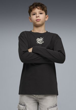 Junge mit verschränkten Armen, der ein schwarzes Puma Street Crew Langarmshirt und hellgraue Cargo-Hosen trägt, vor grauem Hintergrund.